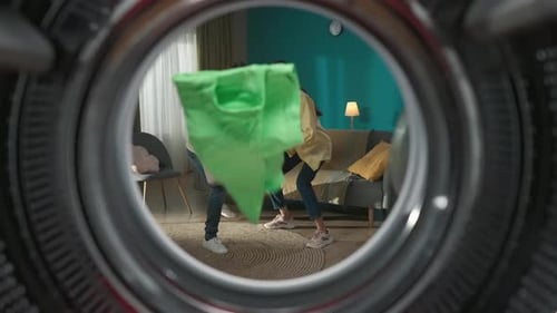 View From Inside the Empty Washing Machine Young Couple with Laundry Basket Throwing Clothes Inside