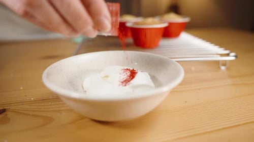 The pastry chef pours red dye into the white cupcake frosting.