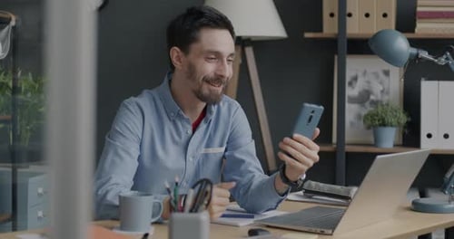 Cheerful Young Man Speaking and Waving Hand Enjoying Video Call with Smartphone From Office