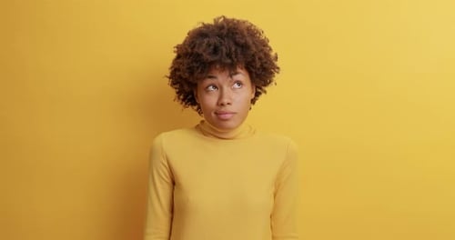 Curly Haired Woman Poses Against Yellow Background Has Hesitant Expression Looks Doubtful As Aske...
