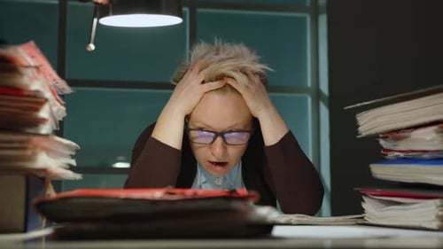 Gestresste Geschäftsfrau schaut sich haufenweise Ordner mit Dokumenten auf dem Tisch an.