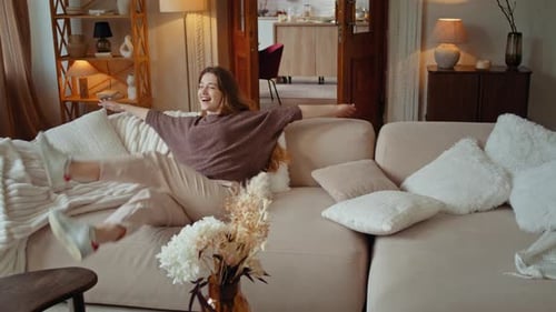 Joyful Young Woman Spins Around and Falls Down Onto Sofa
