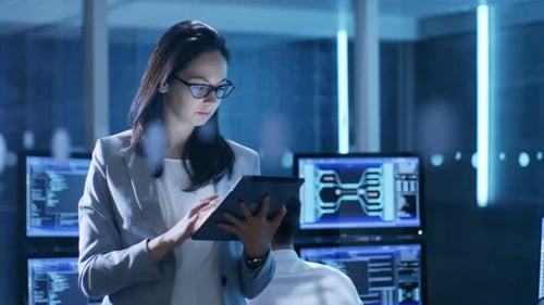 Young Female Government Employee Wearing Glasses Uses Tablet in System Control Center. In the Backg