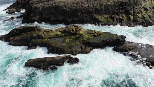 Drone looks down over sea rocks as waves crash into it.