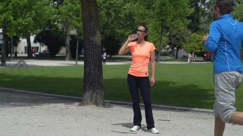 City park joggers man running woman resting and drinking water in super slow motion