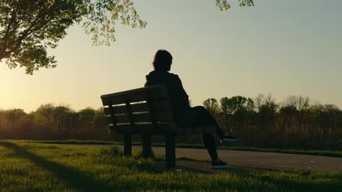 Eine Silhouette einer Frau sitzt auf einer Bank in der Nähe des Sees und ruht sich bei Sonnenuntergang aus