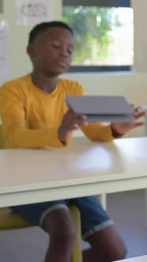 Vertical video: Lifting head, boy in yellow shirt taking gray tablet and viewing lesson in class