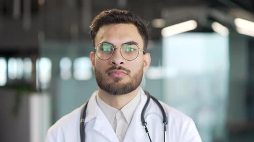 Portrait of serious handsome bearded doctor in white coat standing in modern hospital clinic looking