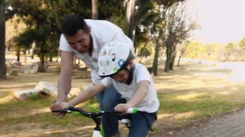 Father teaches son to ride bicycle on park pathway steadicam shot