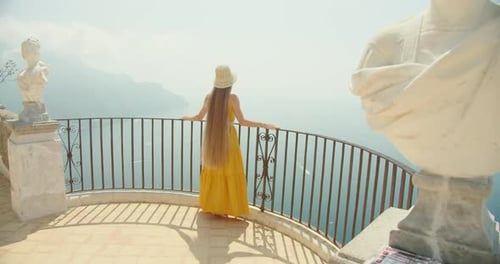 A woman gazes at the sea from a scenic overlook in Italy, with statues and ironwork on the backgroun