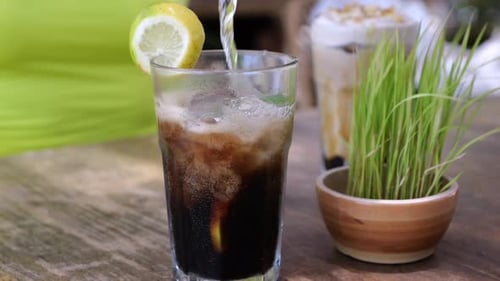 Pouring of Soda Water Into Glass with Ice and Lemon on Outdoor Cafe Table Gas Bubbles Rise to the