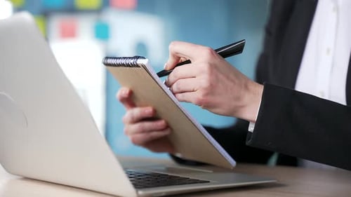 Close up of a woman's hands in office attire taking notes on a notepad while watching a video call