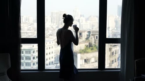 Woman sipping coffee silhouetted admiring morning city view from home window