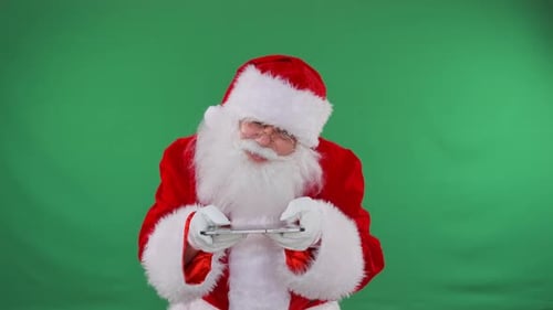 Santa Claus gamer using digital tablet touching screen with fingers standing on green background.