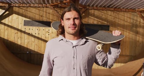 Video of happy caucasian male skateboarder holding skateboard in skate park