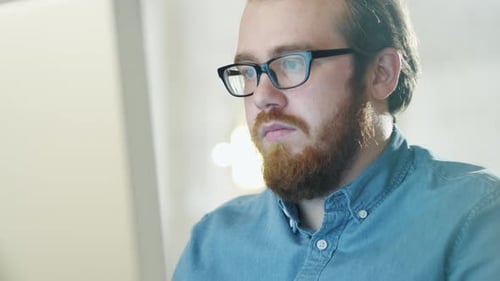 Portrait of a Bearded Young Man Wearing Glasses Sitting in His Office Working on a Computer. Comput