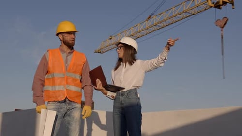 Engineers discussing blueprint on construction site with crane and laptop