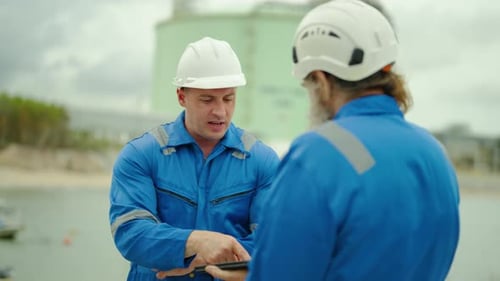 Two caucasian male engineers discuss industrial project plans using a digital tablet at a coastal