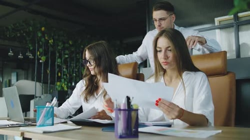 A Manager Stands Beside Two Working Women in a Modern Office Observing Their Progress and Ensuring