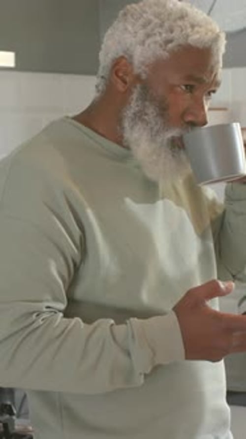 Vertical video: Lifting gray mug, sipping man in sage top using cell inspecting dark jar in kitchen