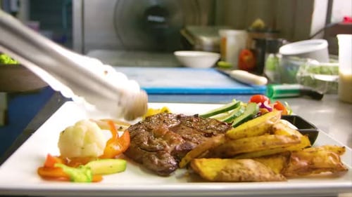 Chef using metal tongs to place grilled vegetables on plate with steak and potatoes