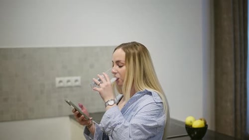 Woman Drinks Water While Using Smartphone in Kitchen