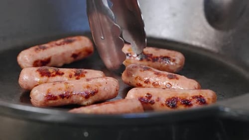 Tongs reach into hot pan of cooked breakfast sausages and grab one with grill marks to plate