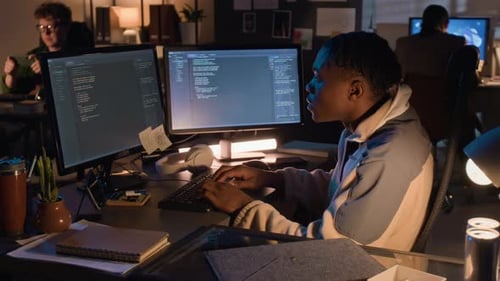 Black Man Coding on Dual Monitors in Office at Night