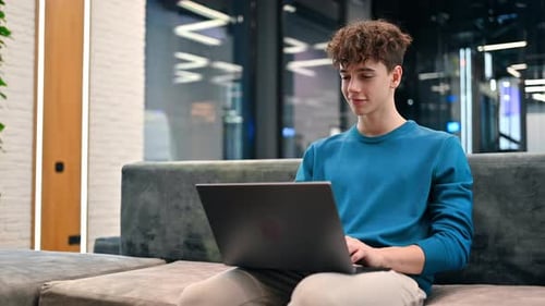 Un joven sonriente que trabaja con una computadora portátil mientras está sentado en un sofá en una oficina