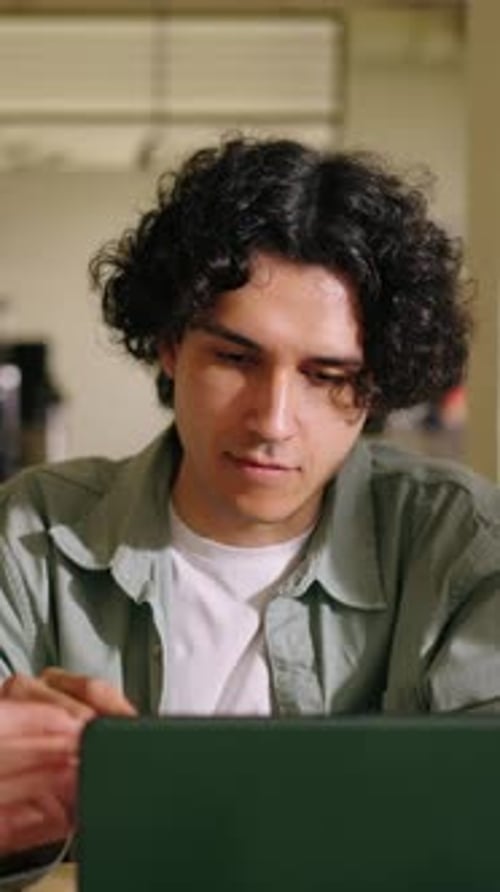 Thoughtful Young Man with Curly Hair Using a Digital Tablet While Studying in a Cozy Cafe Focusing