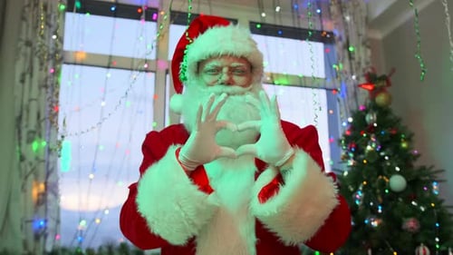 Der Weihnachtsmann bläst Partyhorn in Nahaufnahme, hat Spaß und winkt mit der Hand, vor dem Hintergrund des Weihnachtsbaums