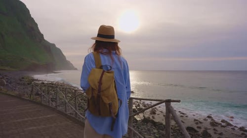 Happy Young Woman Tourist Walks Along the Ocean in a Hat and Backpack on Observation Deck Enjoys Top