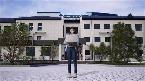 Girl Student Smiling And Typing On A Laptop in Front of a School Building