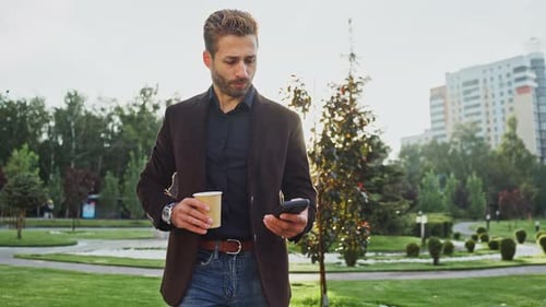 Man Enjoying Coffee While Checking Smartphone in a City Park During Sunset