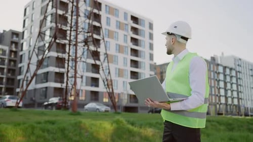 Male Engineer Standing Outside in Protective Gear Inspects Power Line Using Laptop