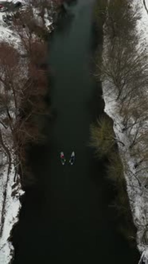 Vertical Video Two Mans Paddleboarding in a Tranquil River with Snowy Trees