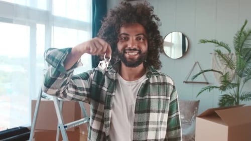 Young Positive Arabian Man Shows Keys with Keychain in Form House Stand in Home