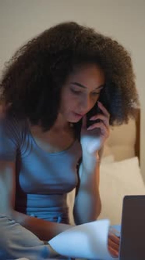 Femme d'affaires afro-américaine furieuse se disputant au téléphone, regardant dans un ordinateur portable et du papier au lit la nuit