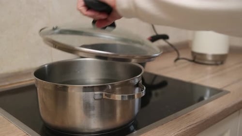 Hand lifting lid from metal pot on an electric stove in a kitchen setting. Indoor cooking concept