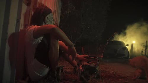 Masked Figure Sits with Chainsaw Next to Cat at Night