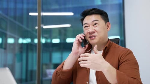 Confident asian businessman talking on mobile phone while sitting at workplace in modern office.