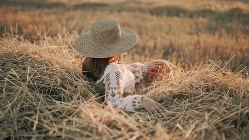 Unrecognizable Woman in Straw Hat and Ethnic Blouse Posing Lying on Hay in Countryside at Sunset