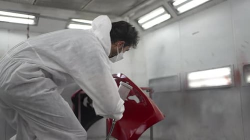 The mechanic is spraying a car bumper at a car repair shop.