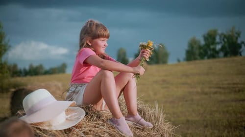Girl Sitting on Hay Bale Playing with Flowers in Farm Field