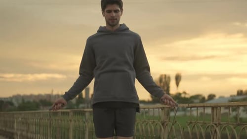 A slim young sportsman is jumping with a rope outside on the bridge in the morning