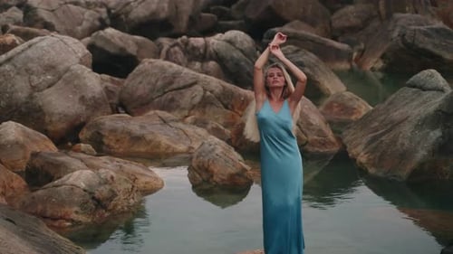 Elegant Woman in Blue Gown Stands Amidst Serene Rocky Waterscape Arms Raised She Embodies