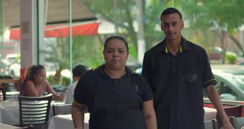 Latina waitress standing next to young male waiter in restaurant, arms relaxed by sides, looking