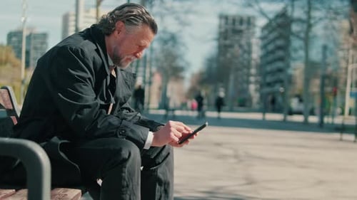 Man on Bench Uses Smartphone in Urban Park