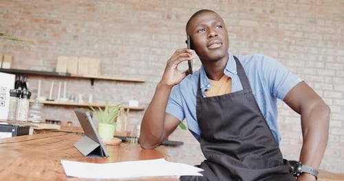African american male barista using tablet and talking on smartphone at coffee shop, slow motion