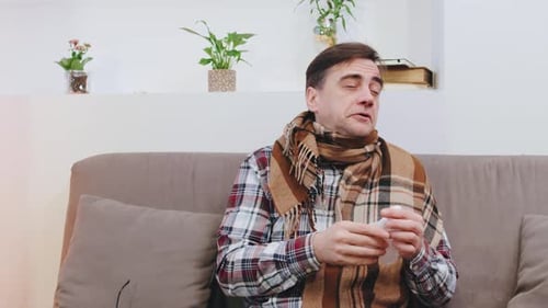 Ein älterer Mann, eingehüllt in eine gemütliche Decke, sitzt auf einem Sofa, umgeben von Taschentüchern, niest, hustet und
